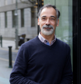 A man with olive skin, brown eyes, dark grey hair and dark grey moustache and beard wearing a navy v-neck jumper over a lavender-coloured shirt smiling at the camera.